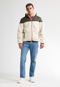 Fleecejack in crème met olijfgroene schouders en capuchon, voorrits en rode accenten. Gecombineerd met lichtblauwe jeans en witte sneakers.