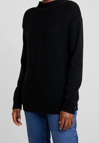 Pull en maille noir avec une surface texturée, col haut et fentes sur les côtés, associé à un jean en denim bleu, mettant en avant une coupe décontractée.