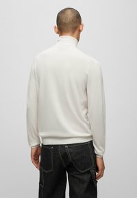 Weißer, langärmeliger Rollkragenpullover mit glatter Textur; verfügt über gerippte Bündchen und Saum; kombiniert mit dunklen Jeans.