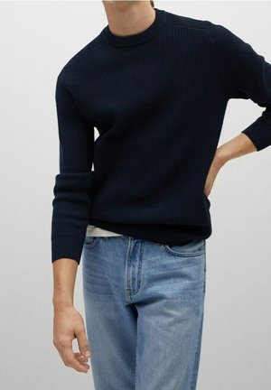 Person trägt einen dunkelblauen, gerippten Pullover mit hochgekrempelten Ärmeln und helle blaue Jeans, steht mit einer Hand in der Hüfte und der anderen entspannt.