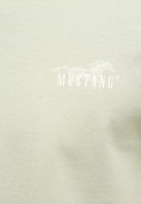Logo Mustang avec un cheval au galop au-dessus du mot « MUSTANG » sur un fond en tissu vert clair.