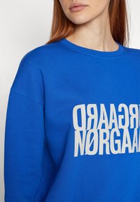 Femme aux cheveux roux portant un sweat-shirt bleu vif avec un texte miroir blanc "NØRGAARD" sur la poitrine.