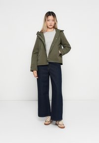Femme portant une veste vert olive, une chemise gris clair, un large jean bleu foncé et des baskets beige, se tenant contre un fond blanc uni.
