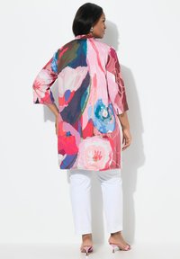 Kolorowy płaszcz z lekkiej tkaniny w kwiatowy wzór, z rękawami do połowy przedramienia, bocznymi rozcięciami i luźnym krojem, zestawiony z białymi spodniami.
