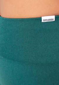 Close-up van de taille van een persoon die een teal gekreukt stof draagt, met een wit label met de tekst "SMILODOX" bevestigd aan de tailleband.