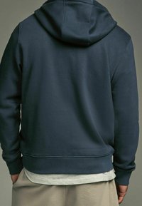 Sweat à capuche bleu marine avec une poche kangourou, des poignets et un ourlet côtelés, une texture douce, une coupe décontractée, et une capuche à l'arrière. Superposé sur une chemise claire.