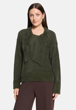 Young woman wearing a dark green fuzzy sweater and dark brown pants, standing against a plain light background.