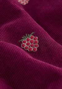 Fuchsiová látka zobrazuje detail malín vyšitých na pozadí s jemnou textúrou, pričom obsahuje červené bobule a zelené listy.