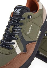 Baskets vert olive avec des accents noirs et marron, dotées d'une tige en daim et en tissu, de lacets contrastants et d'un logo sur la languette.