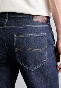 Donkerblauwe denim jeans met contrasterende gele stiksels, voorzien van een mid-rise ontwerp, achterzak met merkembleem en textuurstof.