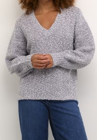 Pull gris avec col en V, motif tricoté texturé et manches lâches. Associé à un jean bleu, mettant en avant un style décontracté.