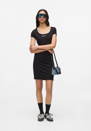 Femeie purtând o rochie neagră cu margini albe, șosete negre, adidași albi, ochelari de soare mari și întunecați, ținând o geantă mică albastră.