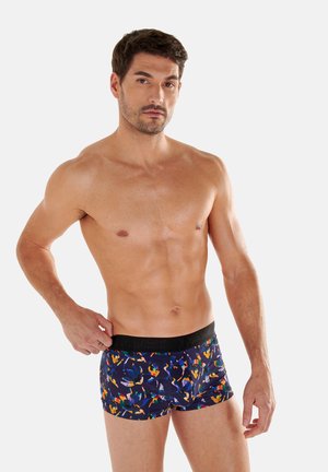 Homme torse nu ajustant un boxer à motif floral multicolore avec ceinture noire sur fond uni.