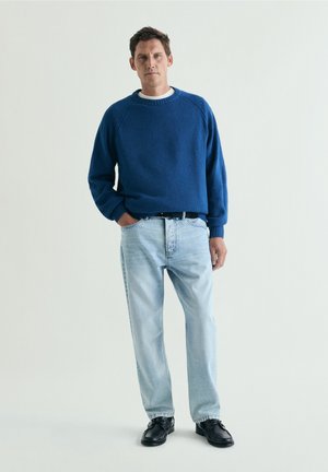 Hombre de pie con las manos en los bolsillos, vistiendo un suéter azul, vaqueros azul claro y zapatos negros contra un fondo liso.