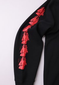 Μαύρο φούτερ με απαλή υφή υφάσματος, διακοσμημένο με επαναλαμβανόμενη κόκκινη γραφική εκτύπωση μιας φιγούρας και τη λέξη "FORMER" κατά μήκος του μανικιού.