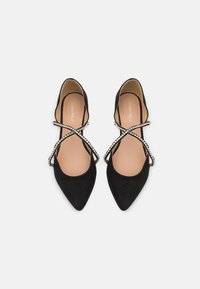Czarne baleriny z ostrym noskiem, z paskami ozdobionymi kryształkami i beżowymi wkładkami na białym jednolitym tle.