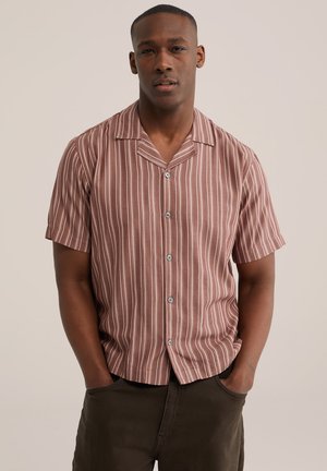 Homme portant une chemise marron à manches courtes, à boutons, avec des rayures blanches verticales, debout les mains dans les poches d'un pantalon foncé, sur un fond uni.