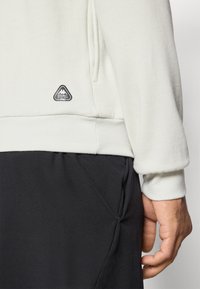 Nærbillede af en person iført en hvid sweatshirt med et lille trekantet logo over taljen og sorte bukser, der viser deres højre hånd.