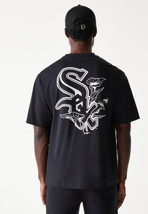 Mand iført sort kasket og sort t-shirt med store hvide stiliserede "Sox" bogstaver og blomstermotiv på ryggen mod ensfarvet baggrund.
