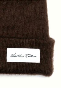 Smeđi pleteni beanie šešir od mekanog materijala, s bijelom etiketom na kojoj piše "Another Cotton" u kurzivnom pismu. Tekstura djeluje udobno i toplo.