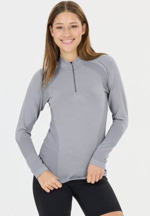 Ung kvinde smilende, iført en langærmet grå sportstop med lynlås foran og sorte tætsiddende shorts, stående mod en ensfarvet baggrund.