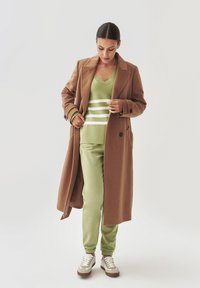 Abrigo de lana marrón sobre un suéter de rayas verdes y joggers verdes. Zapatillas blancas con acentos marrones. Diseño simple y ajustado.