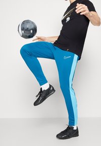 Jogging en turquoise avec une bande bleu clair, associés à un t-shirt noir Nike et des chaussures de sport noires, jonglant avec un ballon de soccer noir et blanc.