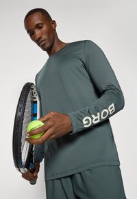 Dlhý rukáv, tmavozelený športový dres s textúrovanou látkou a na rukáve s bielym nápisom "BORG". Drží tenisový míč a raketu.