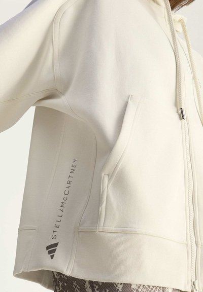 Felpa beige con zip, due tasche, polsini e orlo a coste. Presenta il logo di lato e i lacci sul cappuccio. Tessuto morbido e liscio.