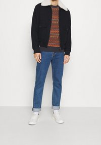 Man i svart jacka med vit fårskinnskrage, mönstrad tröja, uppvikta blå jeans, vita strumpor och vita sneakers som står framför en vit bakgrund.