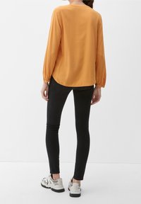 Blouse à manches longues jaune moutarde avec un motif texturé, col rond et poignets froncés, associée à un jean skinny noir et des baskets blanches.