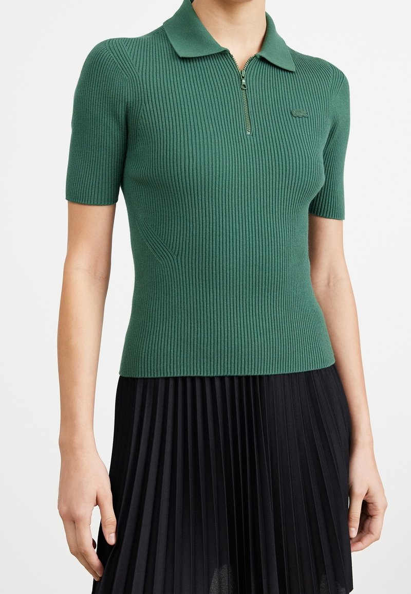 Polo court en tricot côtelé vert avec des manches courtes, doté d'une fermeture éclair quart et d'un logo subtil. Associé à une jupe plissée noire.