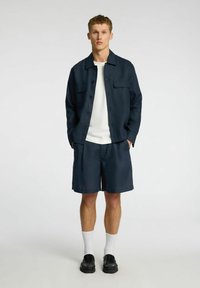 Marineblå kortærmet skjorte med to frontlommer, båret over en hvid T-shirt med rund hals og matchende marineblå shorts, parret med sorte sko.