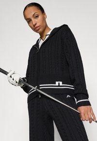 Pulover negru din tricot cu glugă, cu accente albe sub formă de dungi la tiv și un fermoar frontal. Este asortat cu pantaloni potriviți și un club de golf.