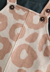 Salopette en imprimé léopard rose avec un tissu texturé, ornée de boutons et de bretelles marron, portée par-dessus une chemise sombre.