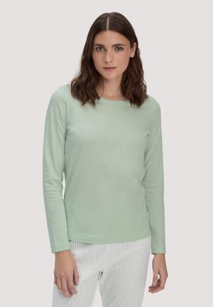 Donna con capelli castani fino alle spalle che indossa una maglia a maniche lunghe verde chiaro e pantaloni bianchi a righe verticali, in piedi davanti a uno sfondo semplice.