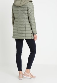 Parka acolchada de color verde oliva con capucha, que presenta un diseño acolchado y botones laterales. Combinada con jeans oscuros y zapatillas deportivas color beige.