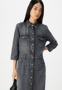 Denimkjole i mørkegrå med krage, trekvartermet, knappestolpe foran, to brystlommer og belte i midjen. Glatt tekstur.