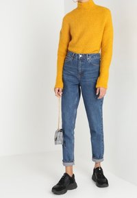 Yellow ribbed knit turtleneck sweater, high-waisted blue denim jeans, black sneakers, and a small checkered handbag with a chain strap.
