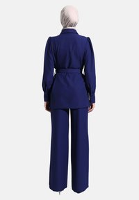 Completo sartorial blu navy con maniche lunghe e vita cintata. Pantaloni a gamba larga completano il design strutturato della giacca. Texture del tessuto liscia.
