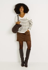 Suéter de cuello en V a rayas crema y oliva, minifalda de ante marrón con botones delanteros, botas altas oscuras hasta la rodilla y bolso marrón con estampado.