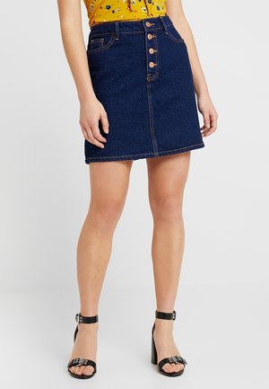 Kvinde iført en mørkeblå denimnederdel med knapper foran, sorte høje hæle med ankelremme og en gul blomsterbluse.