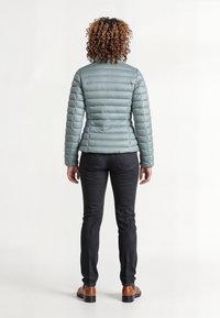 Lichtgroene pufferjack met verticale stiksels, ritssluiting en hoge kraag. Gecombineerd met donkere jeans en bruine schoenen.