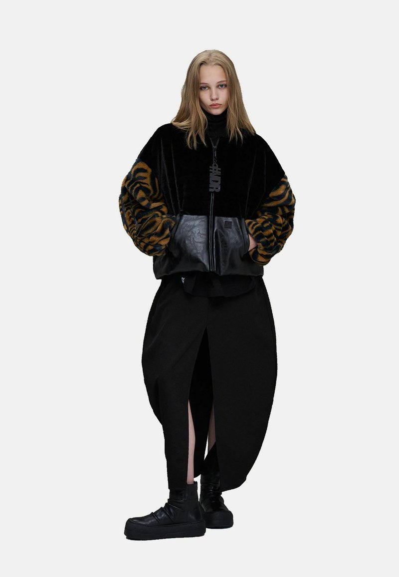 Jeune femme debout portant une veste en fourrure noire et marron à imprimé tigre avec des poches en cuir, une longue jupe noire avec des fentes à l’avant, et des chaussures noires.