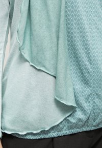 Ljus teal-topp med lagerdesign och textil med mönster underneath, kantad med en rå söm. Slät och mjuk textur genom hela plagget.