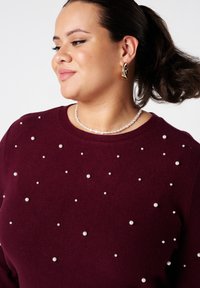 Een vrouw met donker haar dat naar achteren is vastgebonden, draagt een wijnrode trui met verspreide pareldetails, een witte parelketting en sculpturale gouden oorbellen.