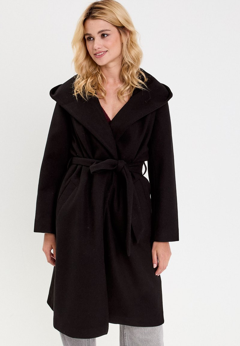 Manteau en laine noire avec un grand col rabattu, une taille ceinturée et des poches latérales, arborant une coupe mi-longue et une texture douce.