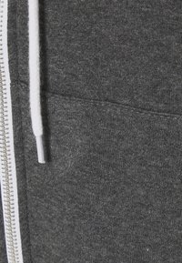 Gros plan sur un sweat à capuche en tissu gris avec un cordon blanc et une fermeture éclair métallique sur le côté gauche.