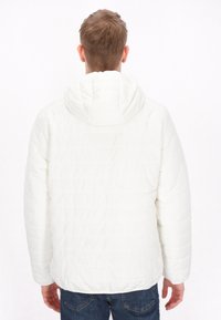 Witte pufferjack met capuchon, met een quilted patroon, lange mouwen en een rechte snit. Draagt over blauwe jeans.