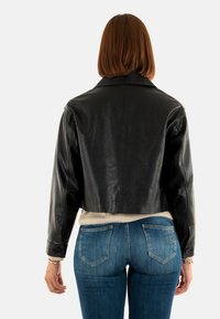 Giacca in pelle nera cropped con una superficie liscia, ampio colletto e polsini con bottone, abbinata a jeans di denim blu e un maglione crema.